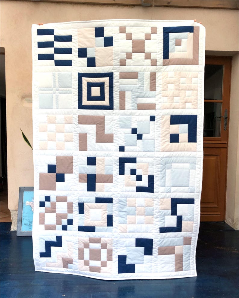 May include: A white quilt with a geometric pattern in navy blue, pale blue, and beige. The quilt has a grid of squares and rectangles, creating a modern, minimalist design. The quilt is displayed against a neutral background.