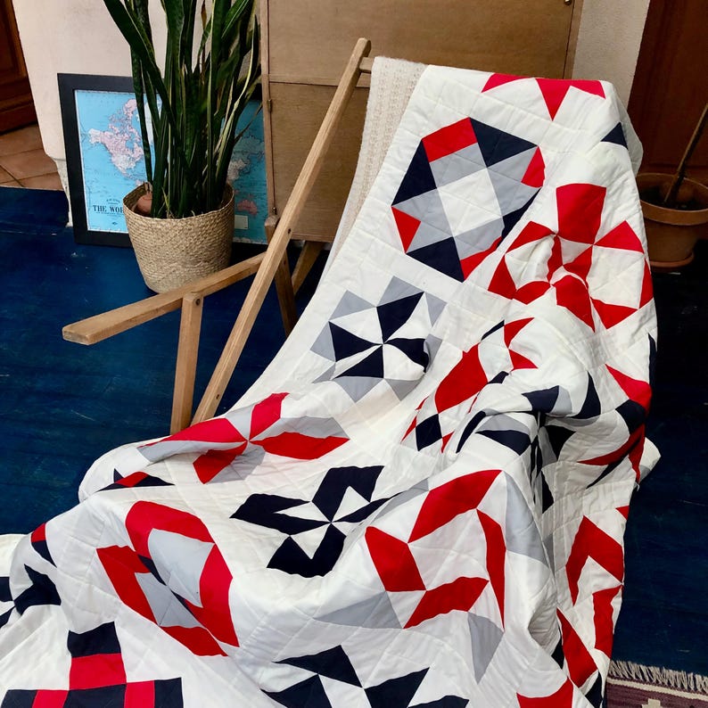 Może przedstawiać: Biała kołdra z czerwonymi, szarymi i czarnymi wzorami geometrycznymi. Kołdra jest złożona i położona na drewnianym krześle.