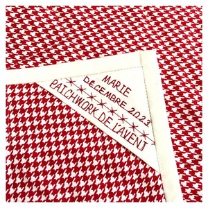 Może przedstawiać: Biały kwadratowy kawałek materiału w czerwono-białą kratę. Na materiale przyszyta jest biała etykieta z wyszytym czerwonym sznurkiem tekstem "Marie D&eacute;cembre 2023 Patchwork de l'Avent".