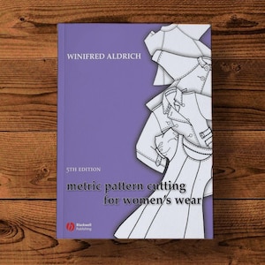 May include: A book titled "metric pattern cutting for women's wear" by Winifred Aldrich, 5th edition, on a purple background. The cover features line drawings of clothing patterns. The book is on a wooden surface.