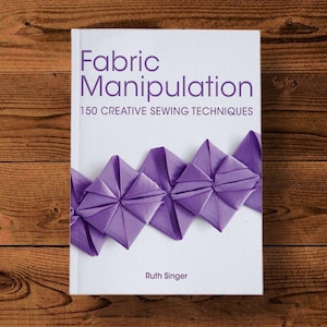 Puede incluir: Portada de libro con el título "Fabric Manipulation: 150 Creative Sewing Techniques" de Ruth Singer. La portada presenta un diseño de tela morada con una serie de cuadrados de tela doblados.