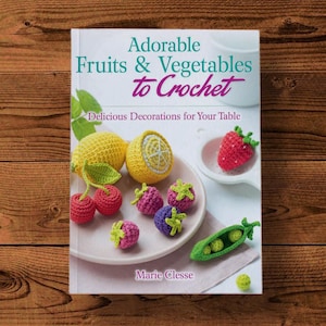 Könnte beinhalten: Buchcover mit gehäkelten Früchten und Gemüse, darunter Zitronen, Kirschen, Erdbeeren, Himbeeren und Erbsen. Der Titel lautet "Adorable Früchte & Gemüse zum Häkeln" mit dem Untertitel "Lecker Dekorationen für Ihren Tisch". Die Autorin ist Marie Clesse.