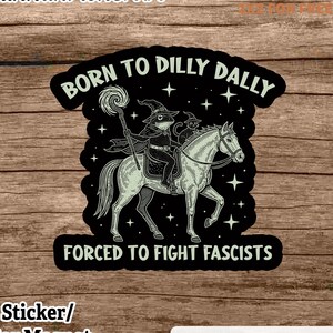 Op de afbeelding: Een zwarte sticker of auto-magneet met de tekst "BORN TO DILLY DALLY" en "FORCED TO FIGHT FASCISTS". De afbeelding toont een kikker en een kat op een paard, beiden met heksenhoeden. Op een achtergrond van houtnerf.