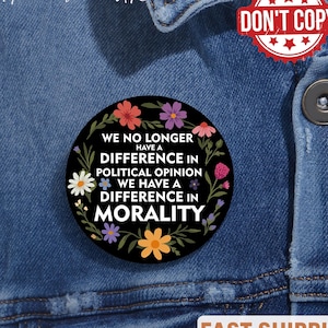 May include: A black button pin with the text "WE NO LONGER HAVE A DIFFERENCE IN POLITICAL OPINION WE HAVE A DIFFERENCE IN MORALITY" surrounded by colorful flowers and leaves. The pin is displayed on a denim jacket.