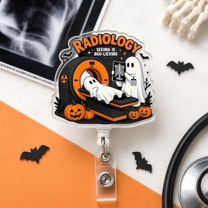 May include: A clear acrylic badge reel featuring a Halloween-themed illustration of two ghosts with a CT scanner, pumpkins, and bats. The text reads RADIOLOGY and SEEING IS BOO-LIEVING. One ghost holds an X-ray showing a skeleton rib cage.