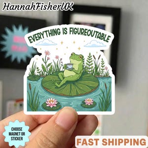 Op de afbeelding: Een sticker met een groene kikker die op een waterlelie zit en een kopje thee vasthoudt. De sticker heeft de tekst "EVERYTHING IS FIGUREOUTABLE" en is omgeven door water, planten en bloemen. De sticker wordt door een hand vastgehouden.
