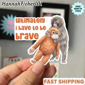 Puede incluir: Una pegatina con una ilustración en acuarela de un mono gris abrazando un peluche naranja. La pegatina tiene el texto "Ultimately I have to be brave" en letras naranjas. La pegatina está sujeta por una mano.
