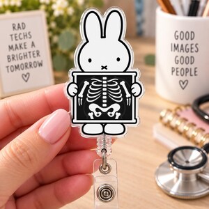 May include: A white acrylic badge reel featuring a cartoon rabbit holding a black and white X-ray image of a human rib cage and spine. The badge reel has a clear plastic strap with a metal snap button and a retractable cord mechanism for ID cards.