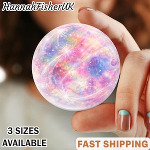 May include: A round, colorful phone grip with a galaxy design in shades of pink, purple, and blue. The phone grip is held in a hand. Text on the image reads "3 SIZES AVAILABLE".