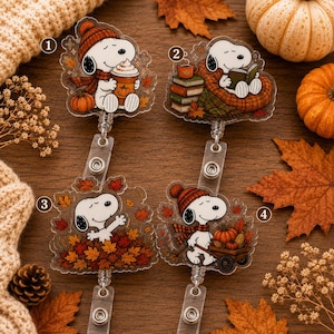 May include: Four acrylic badge reels featuring Snoopy in autumn-themed scenes. Design 1 shows Snoopy with a pumpkin drink, 2 shows Snoopy reading with books, 3 shows Snoopy in a pile of leaves, and 4 shows Snoopy with a pumpkin-filled wooden cart.