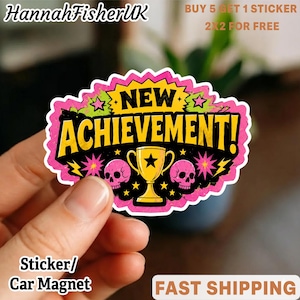 May include: A die-cut sticker featuring a yellow trophy centered between two pink skulls on a black background. The text NEW ACHIEVEMENT! is printed in bold black letters. The design is framed by a jagged pink border with small yellow stars.