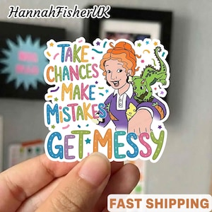 Puede incluir: Una colorida pegatina con una mujer de dibujos animados con pelo rojo y un dragón verde. La pegatina tiene el texto "TAKE CHANCES MAKE MISTAKES GET MESSY" en varios colores. La pegatina está sujeta por una mano.