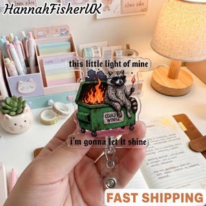 May include: A clear acrylic badge reel featuring a raccoon wearing sunglasses, sitting on a green dumpster filled with orange flames. The text reads, "this little light of mine" at the top and "i'm gonna let it shine" at the bottom, with "COULD BE WORSE" on the dumpster.