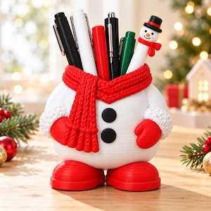 Peut inclure: Un porte-stylo en forme de bonhomme de neige blanc avec une écharpe rouge texturée, des moufles rouges et des bottes rouges. Le bonhomme de neige a deux boutons noirs sur le devant et contient plusieurs stylos, dont un avec un embout bonhomme de neige.