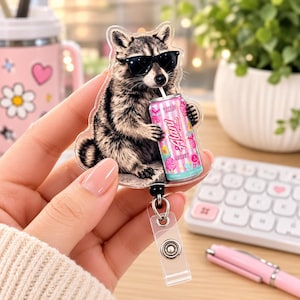 Könnte beinhalten: Ein Ausweishalter aus Acryl mit einer aufgedruckten Illustration eines Waschbären, der eine schwarze Sonnenbrille trägt und aus einer rosa Alani-Energy-Drink-Dose trinkt. Der Waschbär ist grau, schwarz und weiß und hat einen Kunststoffclip.