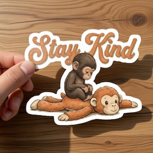 May include: A sticker featuring two cartoon monkeys. One monkey is lying down, and the other is sitting on top. The sticker has the words "Stay Kind" in an orange, retro font.