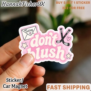 May include: A die-cut sticker featuring the text "don't blush" in a white, rounded font with a pink outline. The design includes a white envelope addressed to "wellsy" with a heart, two crossed black hockey sticks, and small stars.