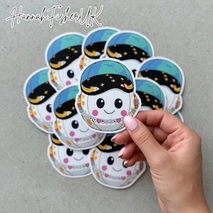 Pode incluir: Uma coleção de autocolantes com uma lua sorridente em estilo cartoon a usar um boné azul e preto com um design de mundo. A lua tem um colar que diz "COPY MOONS". Os autocolantes são brancos com bochechas cor-de-rosa e uma boca preta sorridente.