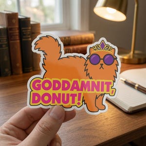 May include: A die-cut sticker featuring an orange, fluffy cat wearing a gold crown with a purple gem and round purple sunglasses. The text GODDAMNIT, DONUT! is printed in bold pink letters with a yellow outline across the bottom of the cat.