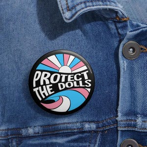 May include: A round black button with the text "PROTECT THE DOLLS" in white. The button features a design with the colors of the transgender flag, including pink, blue, and white, with a sunburst design.