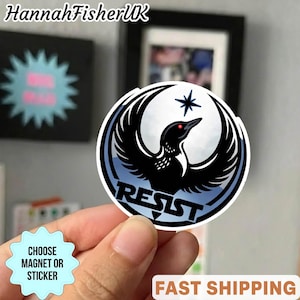 May include: A circular sticker with a black bird design, the word "RESIST", and a star. The bird has a red eye and is set against a blue and white background. The text "CHOOSE MAGNET OR STICKER" is also visible.