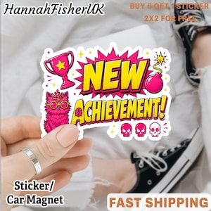 Könnte beinhalten: Ein gestanzter Aufkleber mit dem Text NEW ACHIEVEMENT in Gelb vor einem pinkfarbenen Comic-Hintergrund. Das Design zeigt einen pinken Pokal, eine pinke Katze mit Krone und Brille, eine pinke Bombe und drei kleine pinke Totenköpfe.
