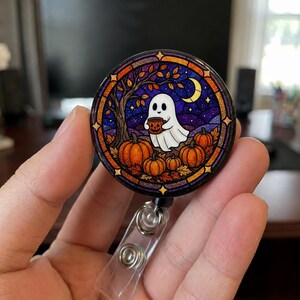 May include: A circular badge reel featuring a stained glass style illustration of a white ghost holding a small pumpkin bucket. The scene includes orange pumpkins, a dark tree, a yellow crescent moon, and a purple starry night sky background.