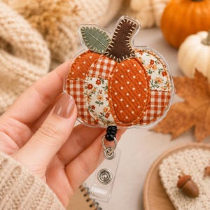 May include: A pumpkin-shaped badge reel featuring a patchwork design of orange, white, and brown fabrics with gingham, polka dot, and floral patterns. The pumpkin has a dark brown stem and a green leaf, all encased in a clear acrylic shell.