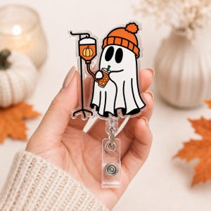 May include: An acrylic badge reel featuring a white ghost wearing an orange beanie. The ghost is connected to an IV drip bag containing an orange pumpkin icon and is holding an orange iced beverage with a green straw. The design has a clear border.