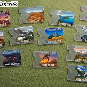 Può includere: Una collezione di portachiavi in acrilico trasparente con viste panoramiche di vari parchi nazionali degli Stati Uniti. Ogni portachiavi mostra un paesaggio diverso, tra cui Arches, Bryce Canyon, Crater Lake e Grand Teton, con il testo "America the Beautiful".