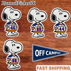Könnte beinhalten: Weiße Snoopy-Aufkleber mit Football-Trikots mit den Namen Tucker, Logan, Graham und Di Laurentis und den Nummern 46, 22, 44 und 66. Ein blaues Wimpel mit dem Text "OFF CAMPUS" ist ebenfalls enthalten.
