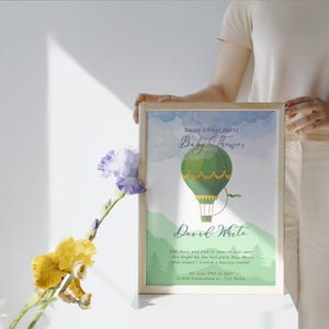 May include: A framed baby shower announcement featuring a green hot air balloon illustration. The text reads "David's First Party Baby Shower" and includes the name "David White" and event details. The frame is held by a person.