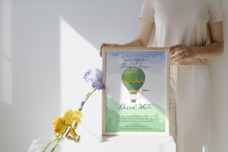 May include: A framed baby shower announcement featuring a green hot air balloon illustration. The text reads "David's First Party Baby Shower" and includes the name "David White" and event details. The frame is held by a person.