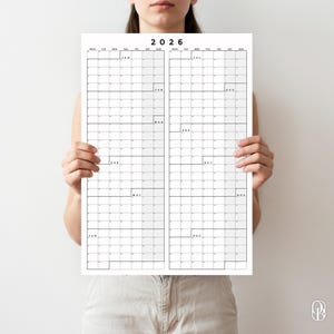 以下が含まれることがあります： ミニマルなデザインの、長方形の2026年カレンダー。カレンダーは月ごとのグリッドに分かれており、各月にラベルが付いています。レイアウトは白黒で、上部に「2026」の年号があります。カレンダーは人が持っています。