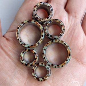 May include: Six brown and tan ear gauges with a speckled pattern are displayed. The gauges are circular with a hollow centre. The speckled pattern is dark brown on a tan background. The gauges vary in size.