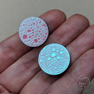 Op de afbeelding: Twee iriserende, ronde plugs met een getextureerd oppervlak. Eén plug heeft een roze en rood patroon, de andere een blauw en turquoise patroon. De plugs worden in een hand gehouden, wat hun unieke, bubbelachtige ontwerp laat zien.