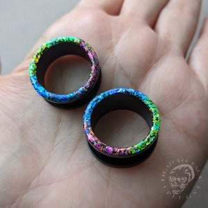 Könnte beinhalten: Ein Paar schwarze Acryl-Ohrenstrecker mit einem regenbogenfarbenen, schimmernden Glitzer-Finish. Die Strecker werden auf einer Hand gezeigt.