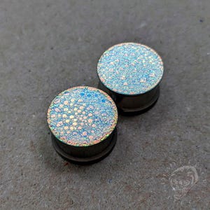 May include: Two iridescent ear plugs with a bubbly, textured surface. The plugs have a dark brown base and a shimmering, light blue and gold top. The plugs are circular and sit on a gray surface.