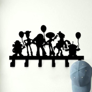 May include: Black metal wall-mounted coat rack featuring a silhouette of the Toy Story characters. The rack has six hooks and is mounted on a white wall. A blue baseball cap and a brown leather handbag are hanging on the hooks.