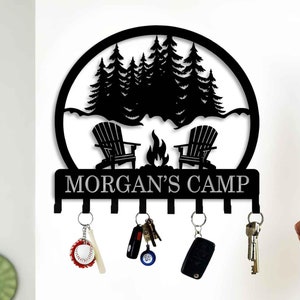 Op de afbeelding: Zwarte metalen sleutelhanger met een silhouet van een kampvuurscène met twee stoelen en bomen. De tekst "MORGAN'S CAMP" staat op de sleutelhanger.