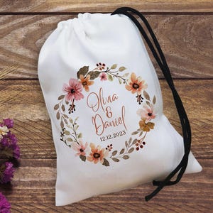 May include: White drawstring bag with a floral wreath design in shades of peach, pink, and brown. The bag features the names "Olivia & Daniel" and the date "12.12.2023" printed in elegant script. The drawstring is black.