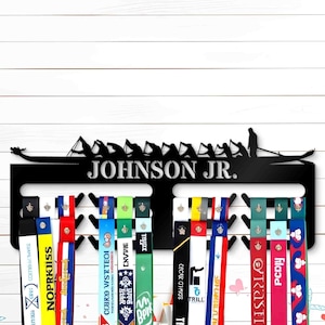 Peut inclure: Un porte-médailles en métal noir en forme de bateau-dragon avec le nom "JOHNSON JR." imprimé sur le devant. Le support est doté de crochets pour accrocher les médailles et les rubans. Le support est monté sur un mur en bois blanc.