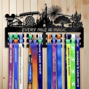 以下が含まれることがあります： テーマパークのシルエットと「EVERY MILE IS MAGIC」の文字が入った黒い金属製のメダルディスプレイ。さまざまな色のリボンを掛けるためのスロットがあり、一部にはテキストが書かれています。