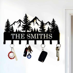 Puede incluir: Perchero de llaves de metal negro con un diseño de silueta de montañas y pinos. El perchero tiene seis ganchos y una placa de nombre personalizada que dice "THE SMITHS".
