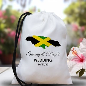May include: White drawstring bag with a black drawstring. The bag features a graphic of the Jamaican flag in the shape of the island of Jamaica. The text "Sammy & Taryn's Wedding 10/27/23" is printed below the graphic.