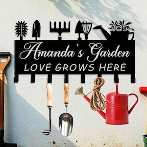 Könnte beinhalten: Schwarzer Gartenwerkzeugständer aus Metall mit Haken. Der Ständer hat ein Schild mit der Aufschrift "Amanda's Garden Love Grows Here".