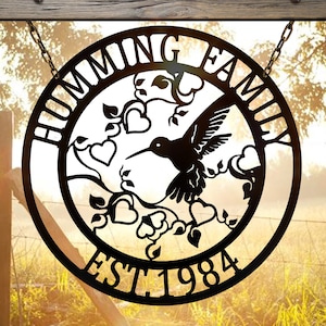 Puede incluir: Letrero circular de metal negro con las palabras "HUMMING FAMILY" y "EST 1984". Un colibrí está en el centro, rodeado de corazones y vides. El letrero cuelga de cadenas. Un artículo decorativo para el hogar.