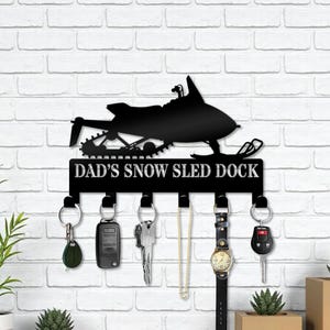 May include: Black metal key holder featuring a snowmobile silhouette and the text "DAD'S SNOW SLED DOCK." The key holder has five hooks with keys, a watch, and other items hanging from them. The key holder is mounted on a white brick wall.