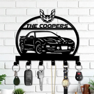 May include: Black metal key holder with a car silhouette, the text "THE COOPERS", and a decorative bird emblem. Includes five key hooks with keys, a watch, and a car remote hanging from them. Mounted on a white brick wall.