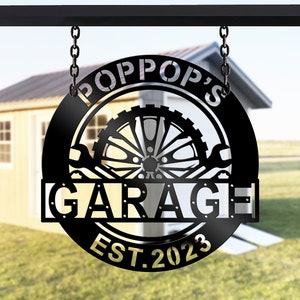 Op de afbeelding: Zwart metalen bord met de tekst "Poppop's Garage Est. 2023" in witte letters. Het bord heeft een gestileerde afbeelding van een band en een moersleutel.